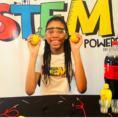 Alaina Davis, Co-Owner and CEO of STEM Powered Kits, smiling while holding lemons during a Fruit Battery STEM experiment, surrounded by STEM tools and equipment.