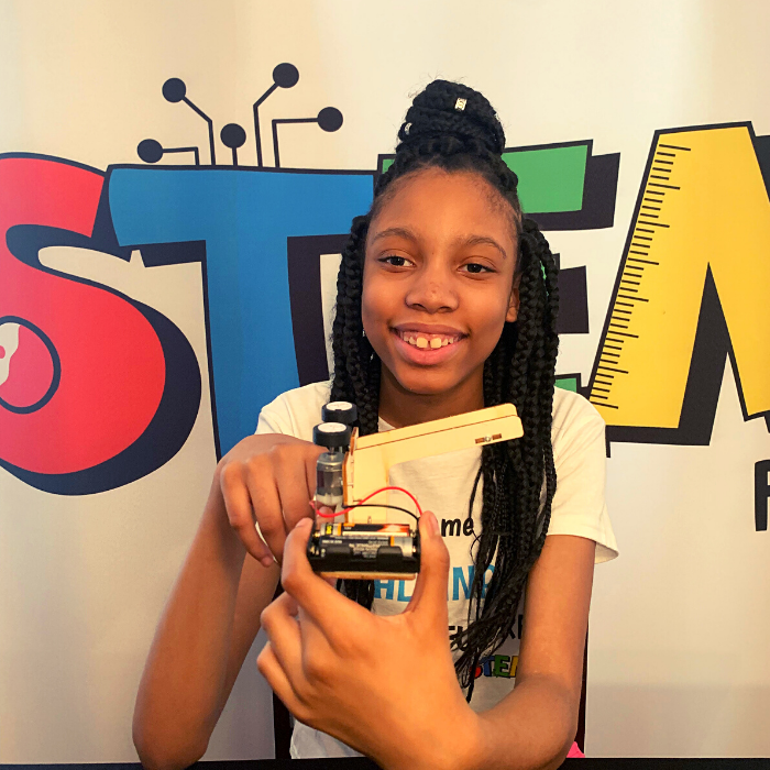 Alaina Davis, Co-Owner and CEO of STEM Powered Kits, holding the assembled Ball Pitching Machine STEM Kit in front of the STEM Powered Kits logo.